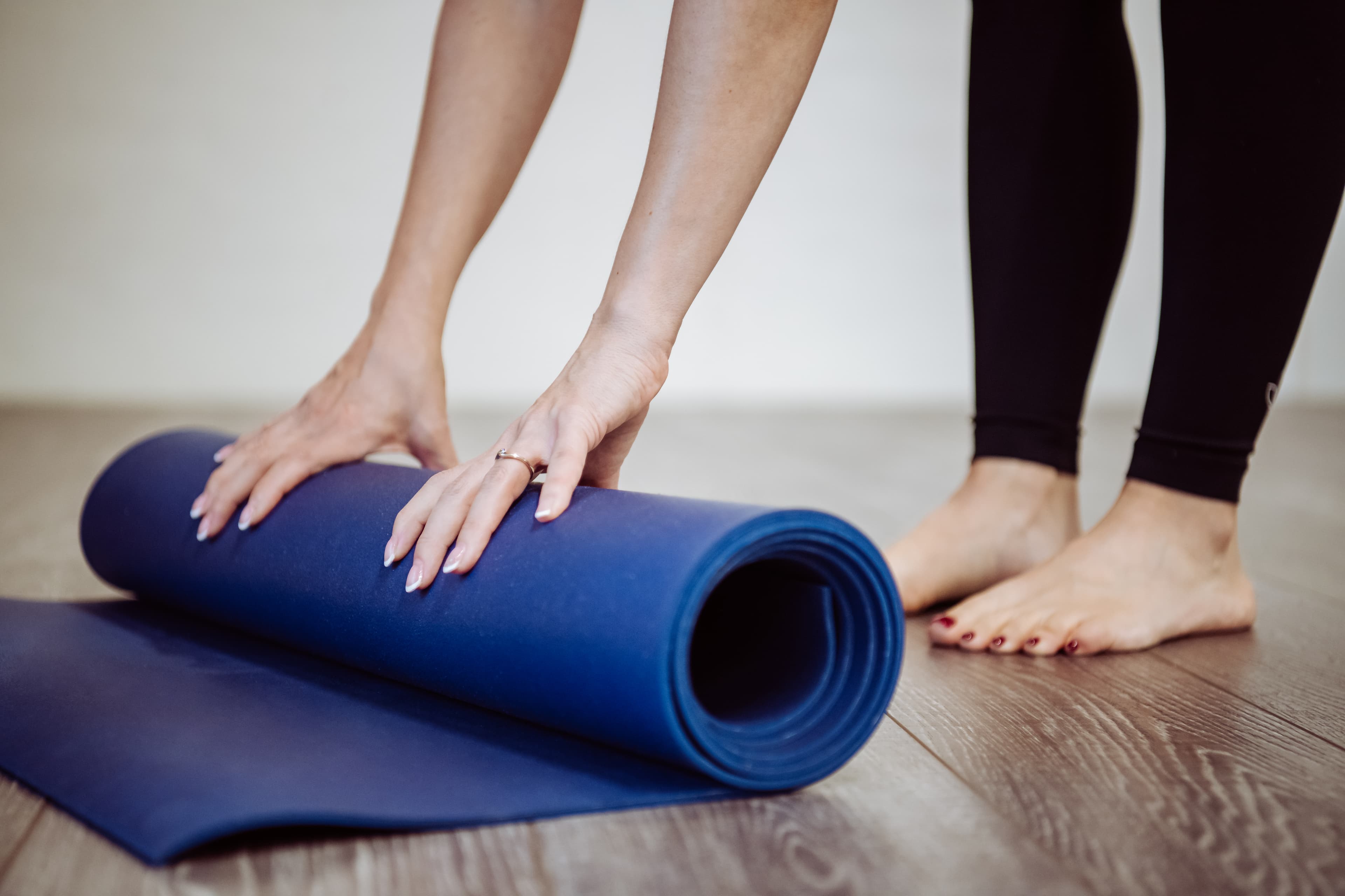 Hände rollen eine blaue Yogamatte auf einem Holzboden neben nackten Füßen sorgfältig zusammen.